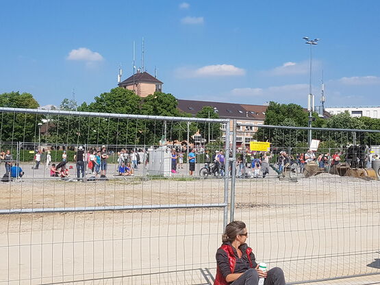 Frau mit Termoskanne sitzt allein vor dem Grenzzaun. Im Hintergrund Demonstranten.