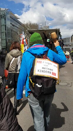 Frau mit Rassel und Plakat auf dem Rücken.