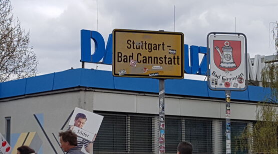 Zwei Ortsschilder von Bad Cannstatt, eines mit dem traditionellen Kannen‑Symbol, im Vordergrund Demonstrierende mit Plakaten
